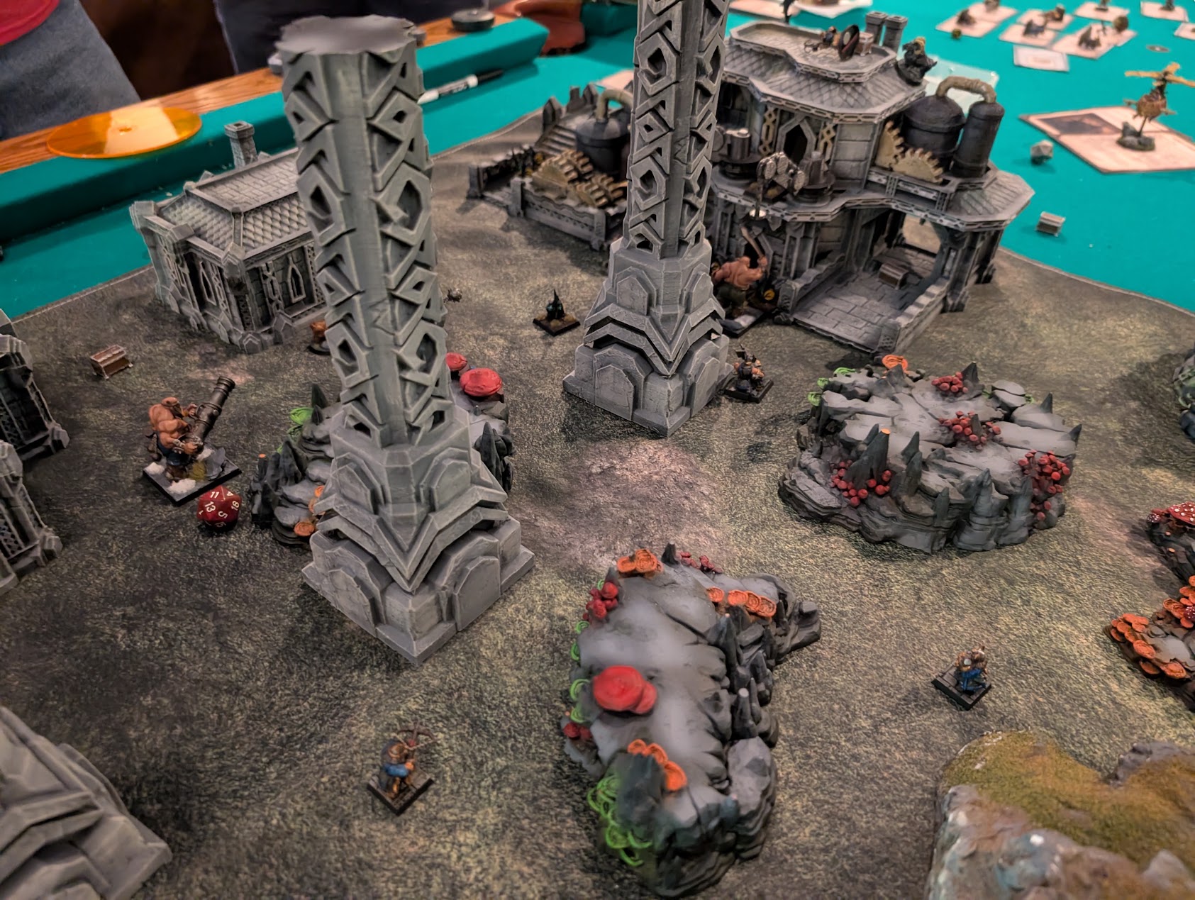 Miniatures and red markers surround towering stone pillars and industrial buildings on the gyrocopter factory battlefield.